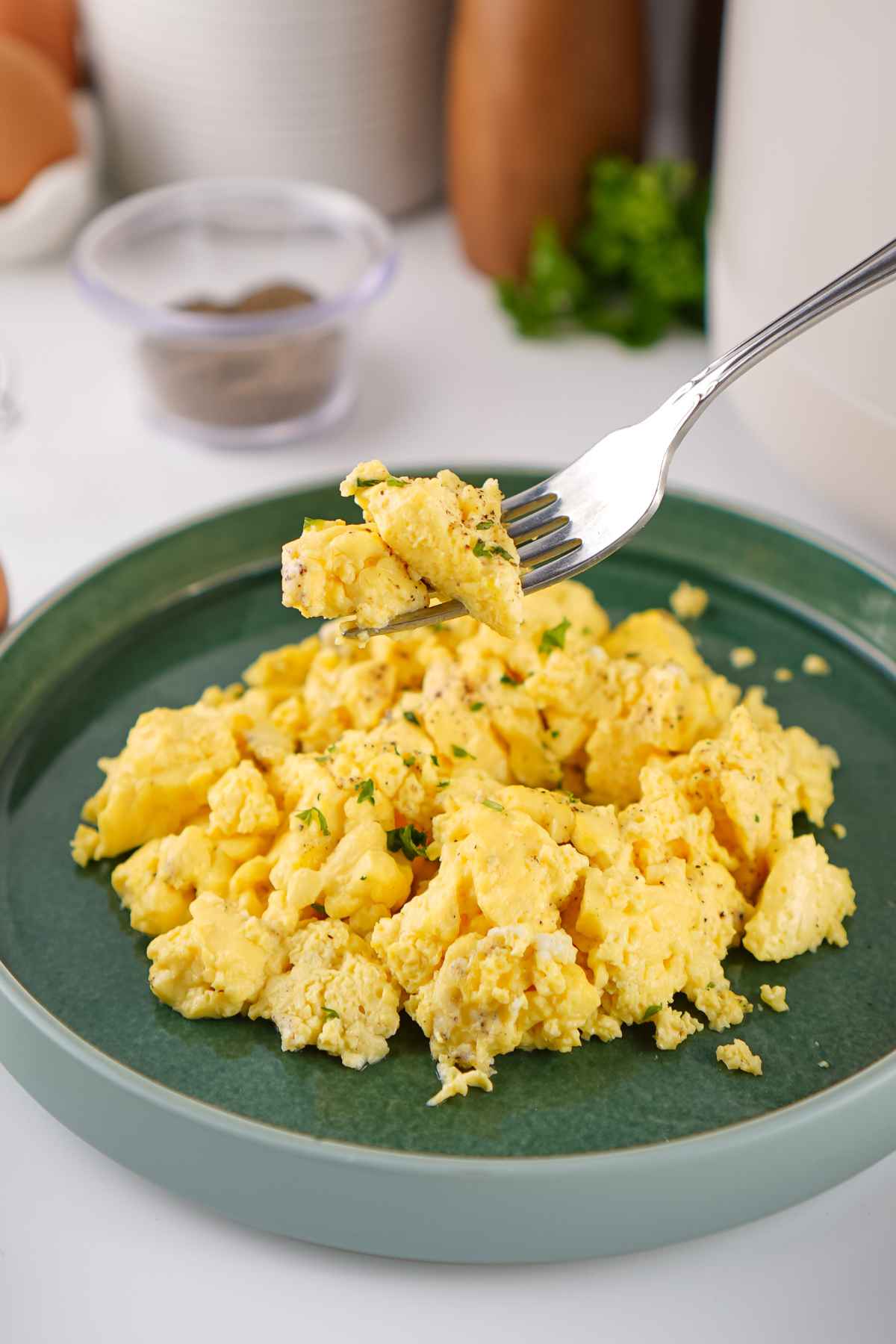 A silver fork takes a bite from a heap of fluffy, seasoned crockpot scrambled eggs served on a dark green plate.