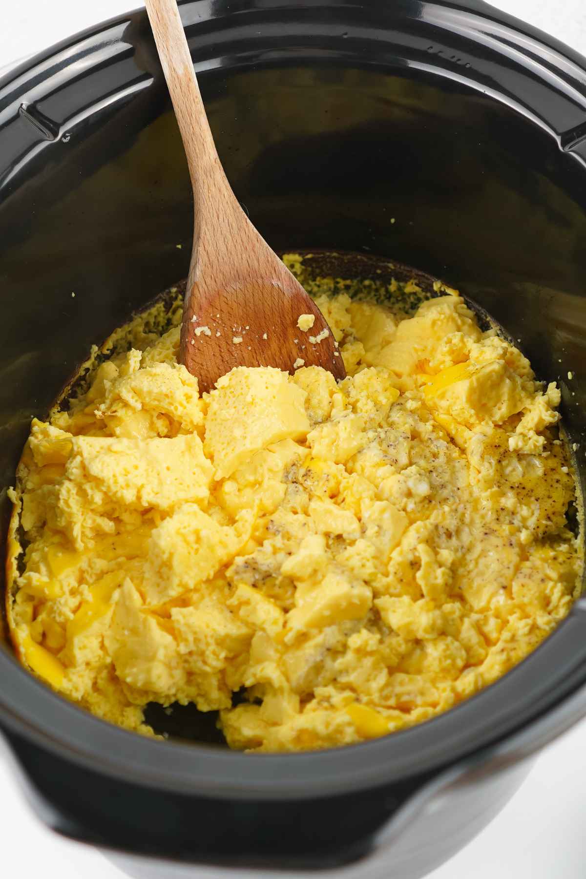 A wooden spoon stirs a large batch of fluffy, yellow scrambled eggs inside a black slow cooker.