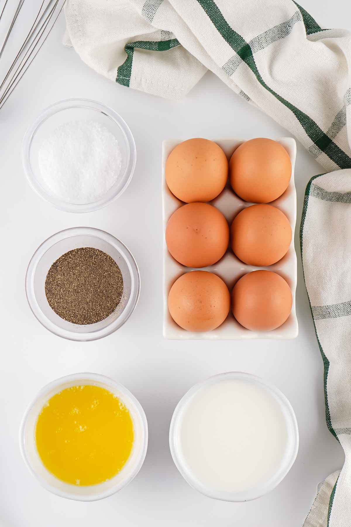 A flat-lay view of ingredients for crockpot scrambled eggs including brown eggs in a ceramic carton, bowls of melted butter and milk, salt, pepper, and a whisk.