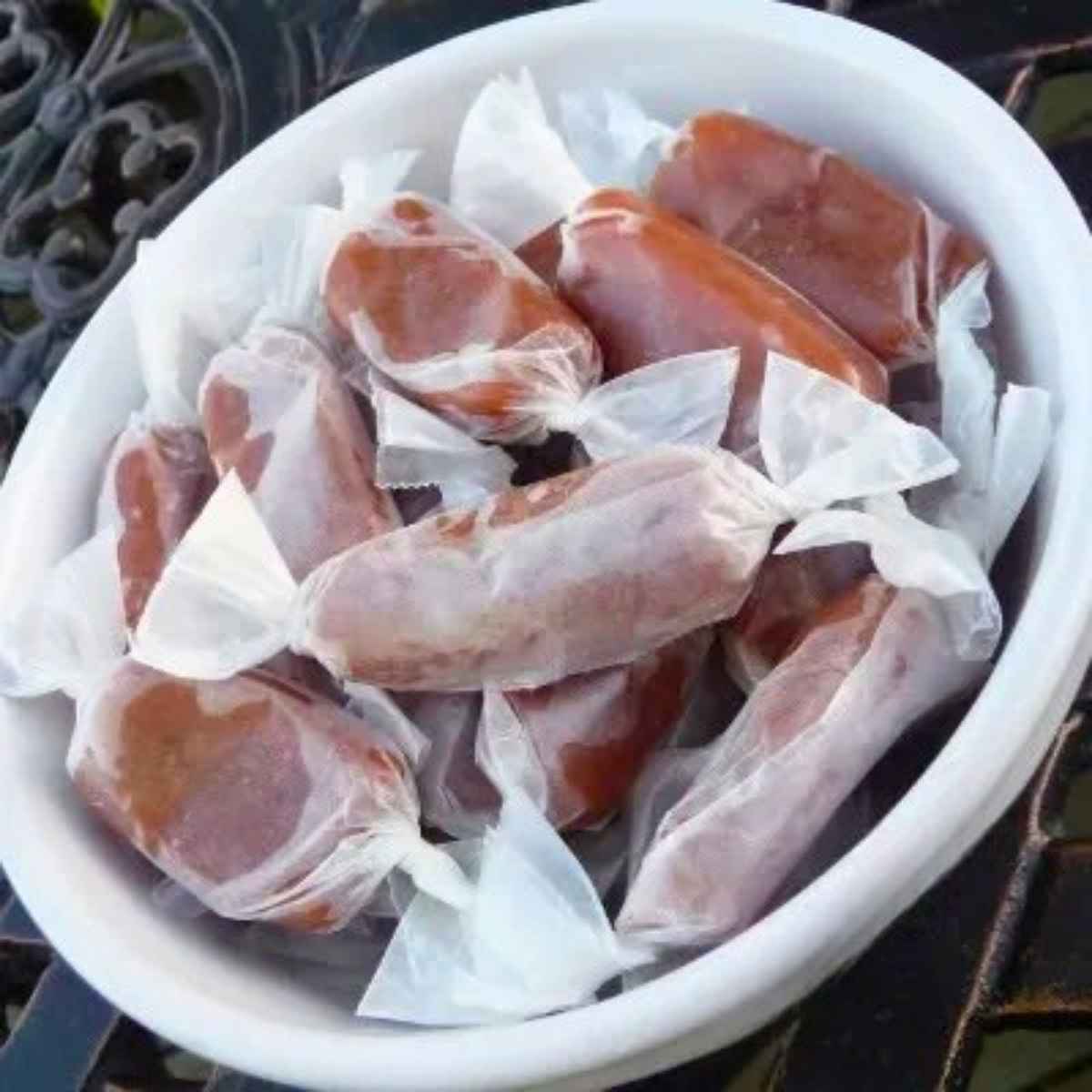 A white bowl filled with several homemade salted caramels individually wrapped in clear wax paper and stacked together.