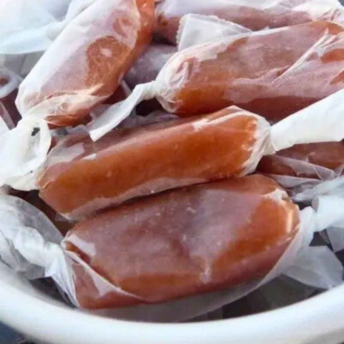 A close up of several homemade salted caramels individually wrapped in clear wax paper and stacked in a white bowl.