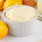 A white fluted ramekin filled with thick, creamy homemade mayonnaise, sitting next to fresh lemons and eggs.