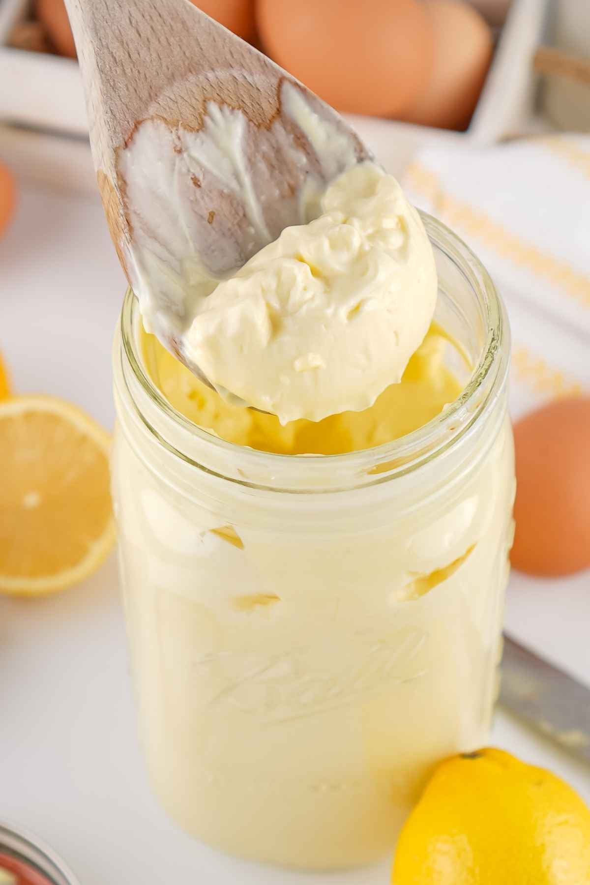 A wooden spoon scooping thick, creamy Homemade Mayonnaise from a glass mason jar, with lemons and eggs in the background.