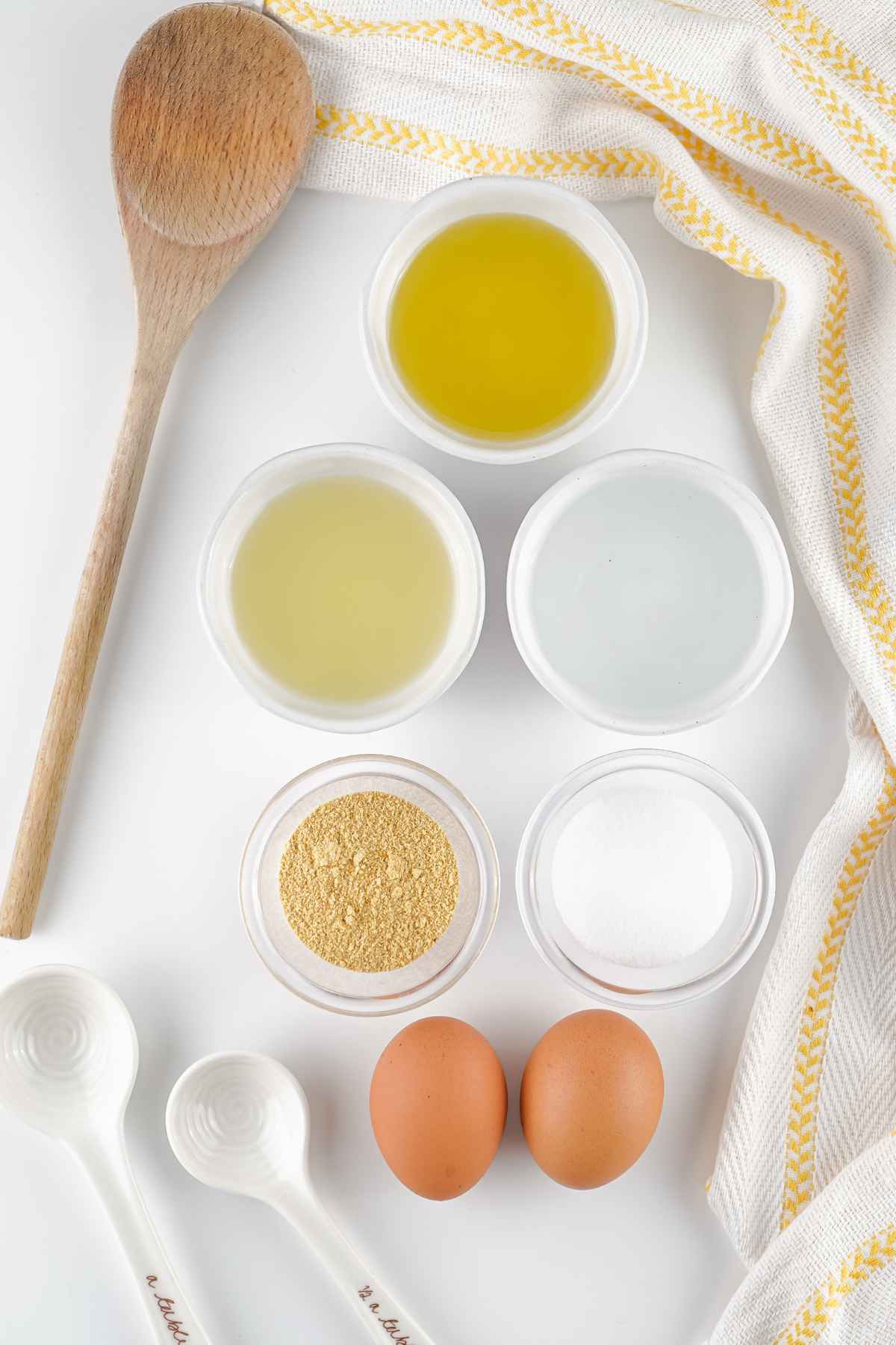 Overhead view of ingredients in small glass bowls including oil, vinegar, mustard powder, and salt alongside two brown eggs and white measuring spoons on a light surface.