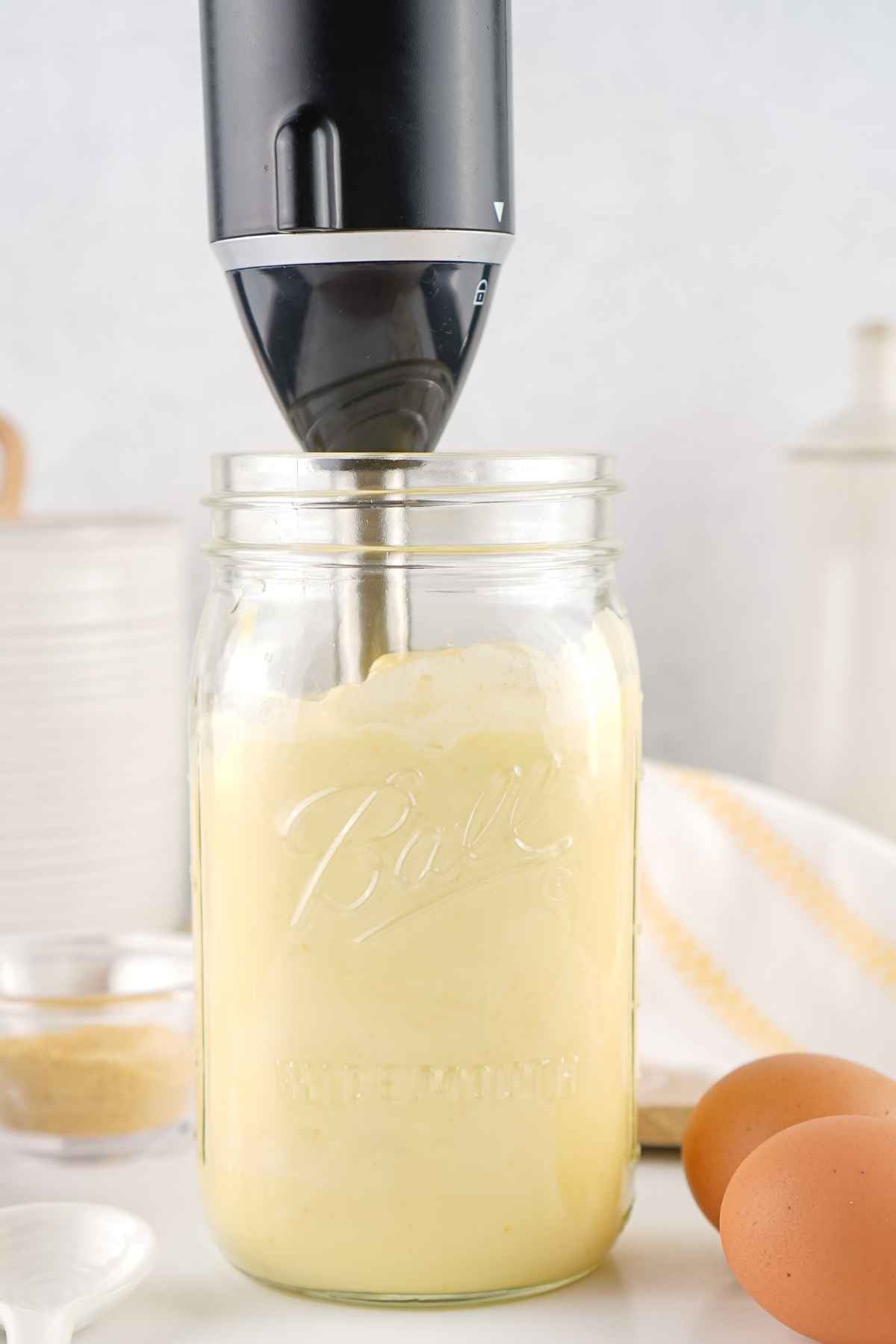 An immersion blender being used to mix a creamy white condiment inside a glass mason jar.