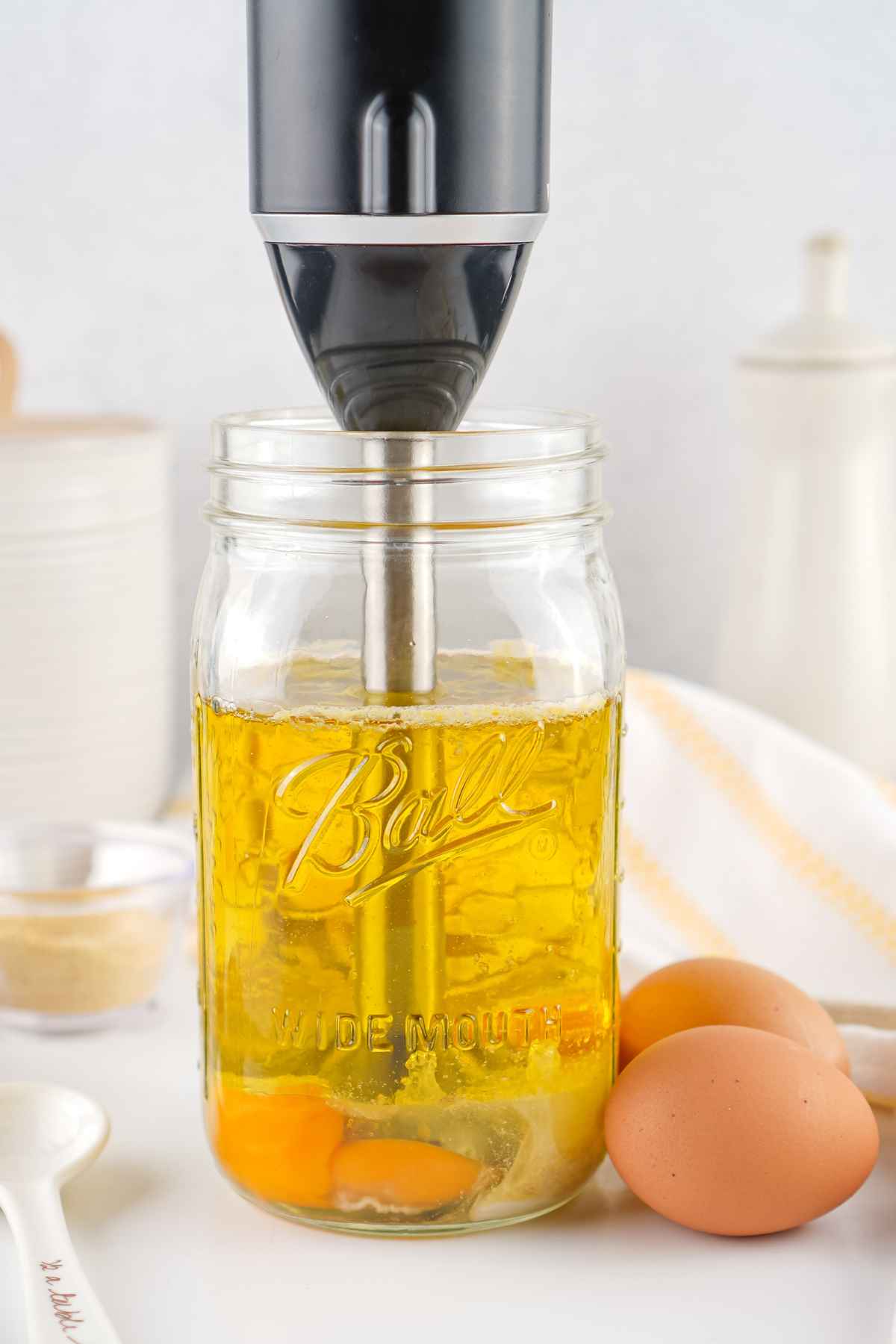 Immersion blender placed inside a glass mason jar filled with oil and eggs on a white countertop.