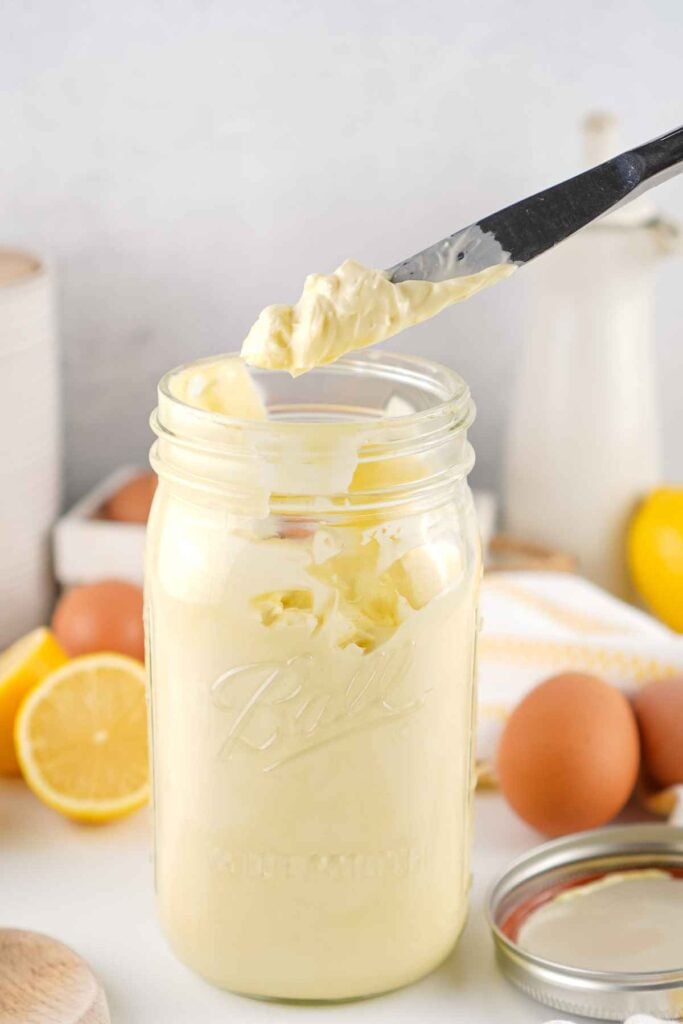 A dollop of thick, creamy mayonnaise spread on the tip of a silver butter knife held over a glass mason jar.