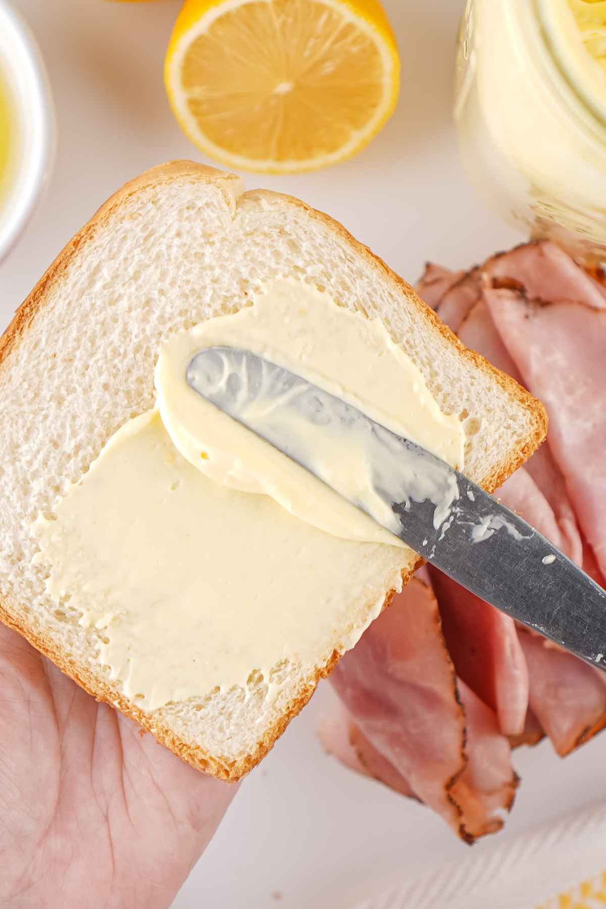A silver knife spreading a thick, creamy pale yellow condiment onto a slice of white bread with ham in the background.