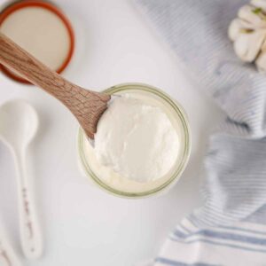 Thick and creamy homemade sour cream on a wooden spoon, held above a glass jar in a bright kitchen setting.