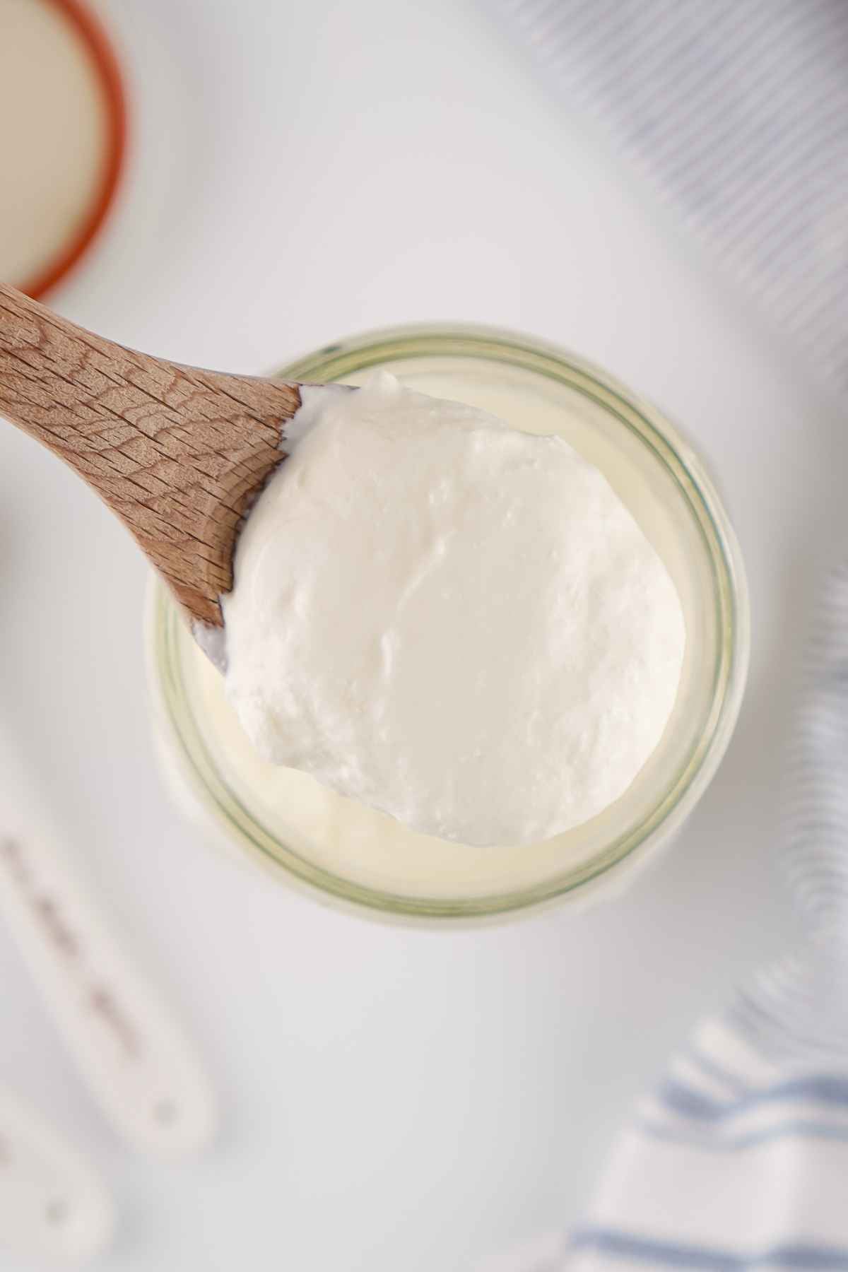 Thick and creamy homemade sour cream on a wooden spoon, held above a glass jar in a bright kitchen setting.
