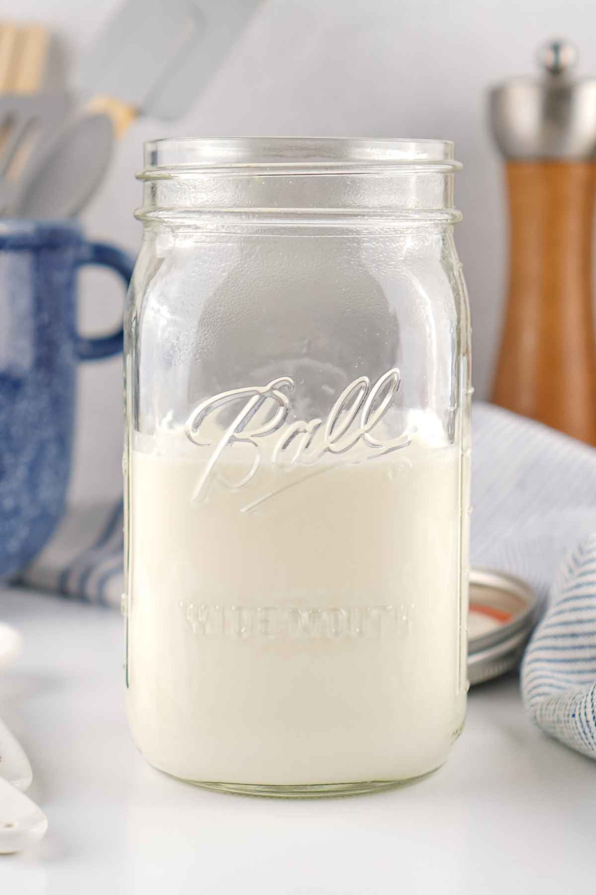 A wide-mouth glass Mason jar filled with a thick, smooth white cultured dairy cream, set on a light surface with a blue patterned mug and kitchen canisters blurred in the background.