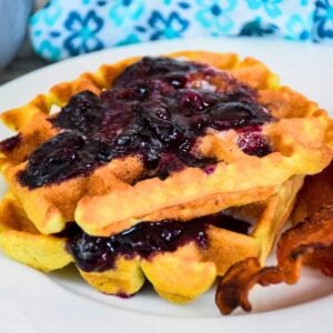 Two stacked keto waffles topped with sugar-free blueberry sauce and a side of crispy bacon.