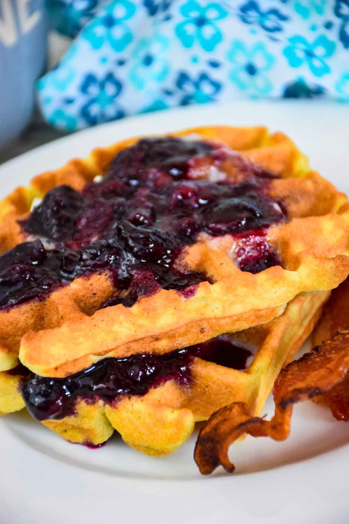 Side view of two keto waffles topped with blueberry sauce on a white plate with a side of crispy bacon.