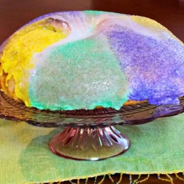 A homemade Mardi Gras King Cake topped with white icing and traditional purple, green, and yellow sanding sugar, displayed on a purple glass cake stand.