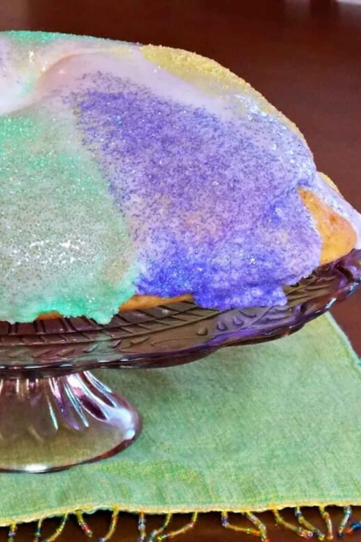 A homemade Mardi Gras King Cake topped with white icing and traditional purple, green, and yellow sanding sugar, displayed on a purple glass cake stand.