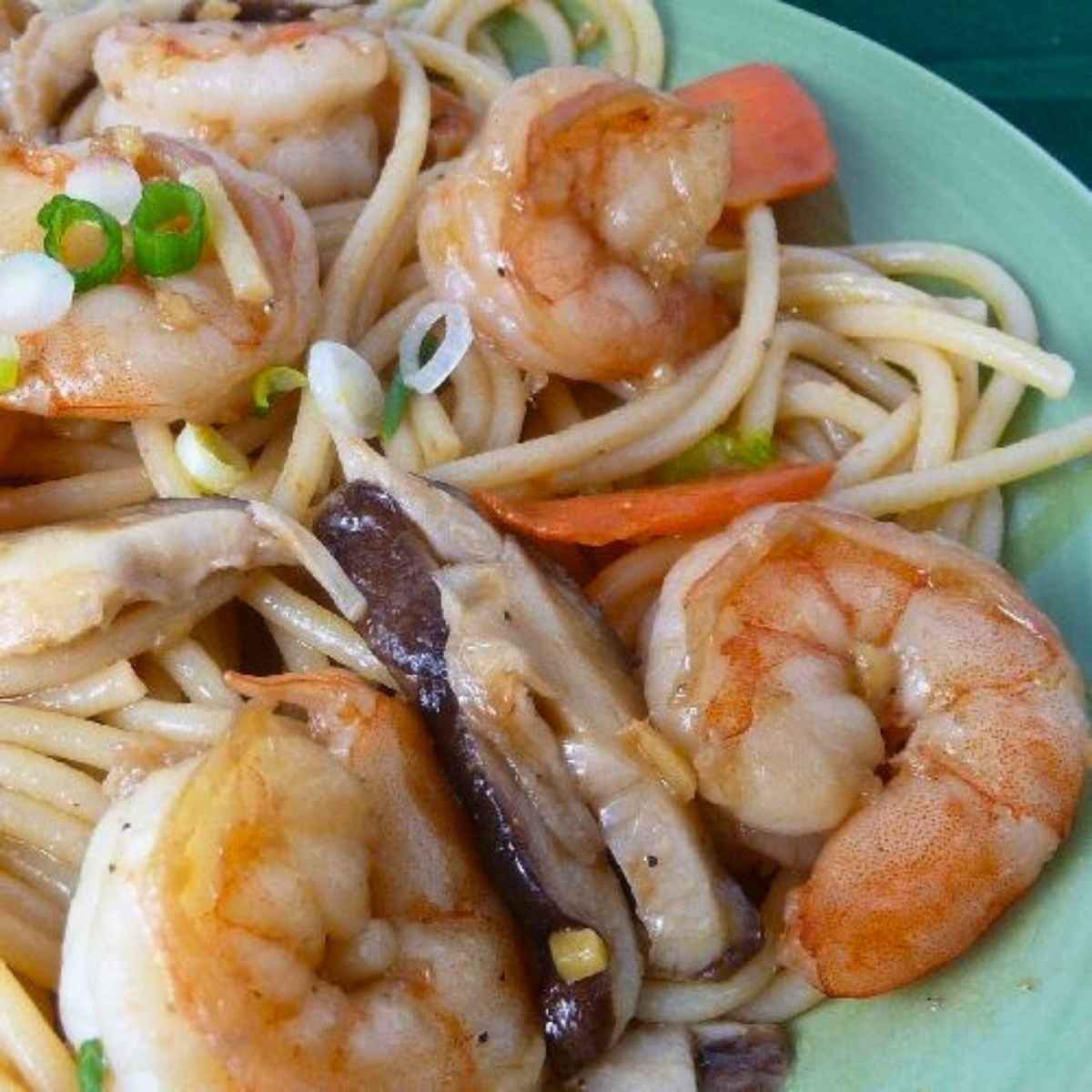 Close up of cooked noodles featuring stir-fried shrimp, sliced mushrooms, carrots, and green onions in a savory sauce.
