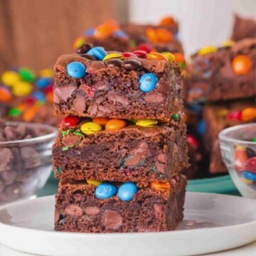 A stach of 3 fudgy cake mix brownies studded with colorful M&M candies on a white plate with more brownies blurred in the background.