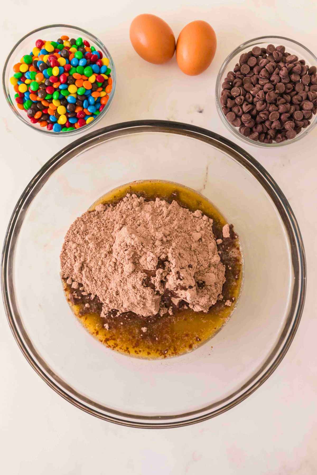 A glass mixing bowl filled with dry mix base, fresh eggs, semi sweet chocolate chips, and bright candy coated chocolates.