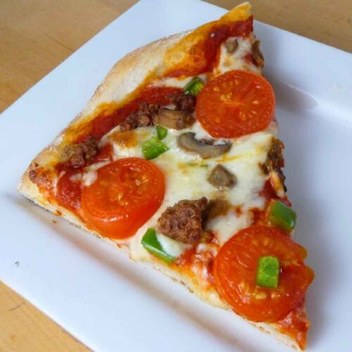 A close-up slice of homemade pizza on a white plate, featuring a thick golden crust topped with melted cheese, tomato slices, green peppers, mushrooms, and sausage.