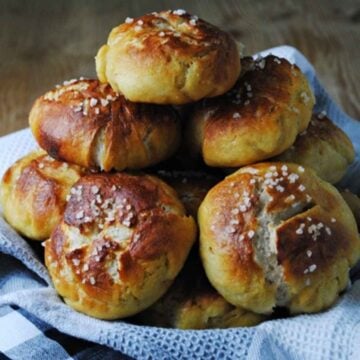 A stack of golden brown homemade pretzel rolls topped with coarse sea salt, resting on a blue and white patterned kitchen towel.