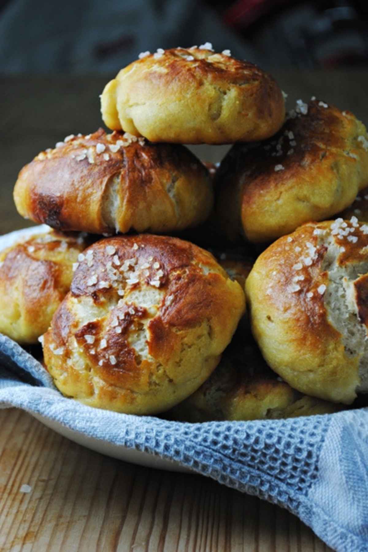 A stack of golden brown homemade pretzel rolls topped with coarse sea salt, resting on a blue and white patterned kitchen towel.