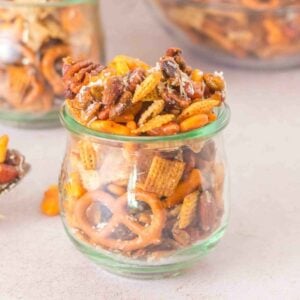 Featured image of homemade Sweet and Salty Chex Mix in a glass jar, showcasing the mix of crunchy cereal, salty pretzels, nuts, and a savory shredded cheese.