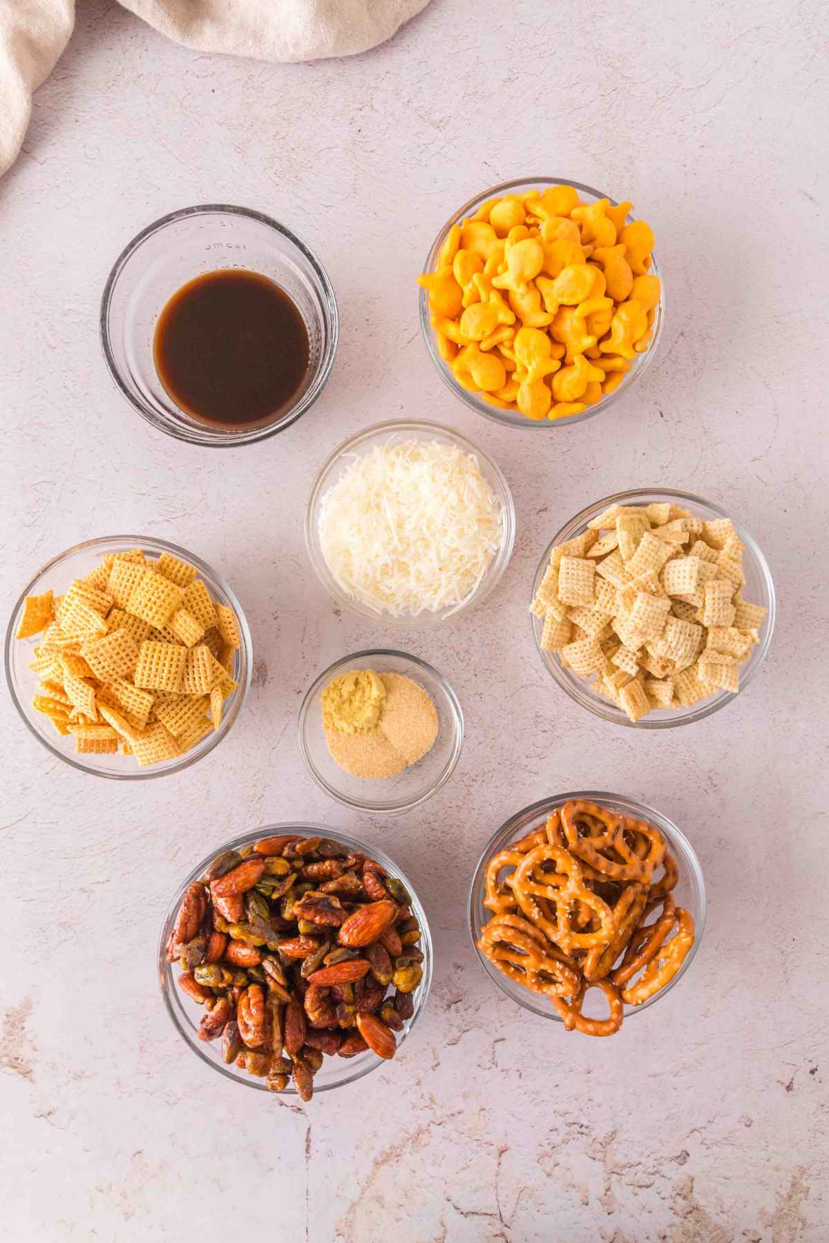 Top down view of six glass bowls containing snack mix ingredients: Chex cereal, mini pretzel twists, goldfish crackers, shredded coconut, mixed nuts, and a small bowl of seasoned butter sauce, all arranged on a light colored stone surface.