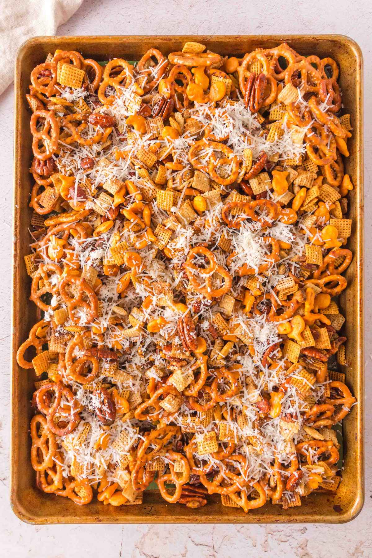 Freshly baked Sweet and Salty Chex Mix on a baking sheet topped with shredded parmesan cheese, including pretzels, nuts, and cereal squares.
