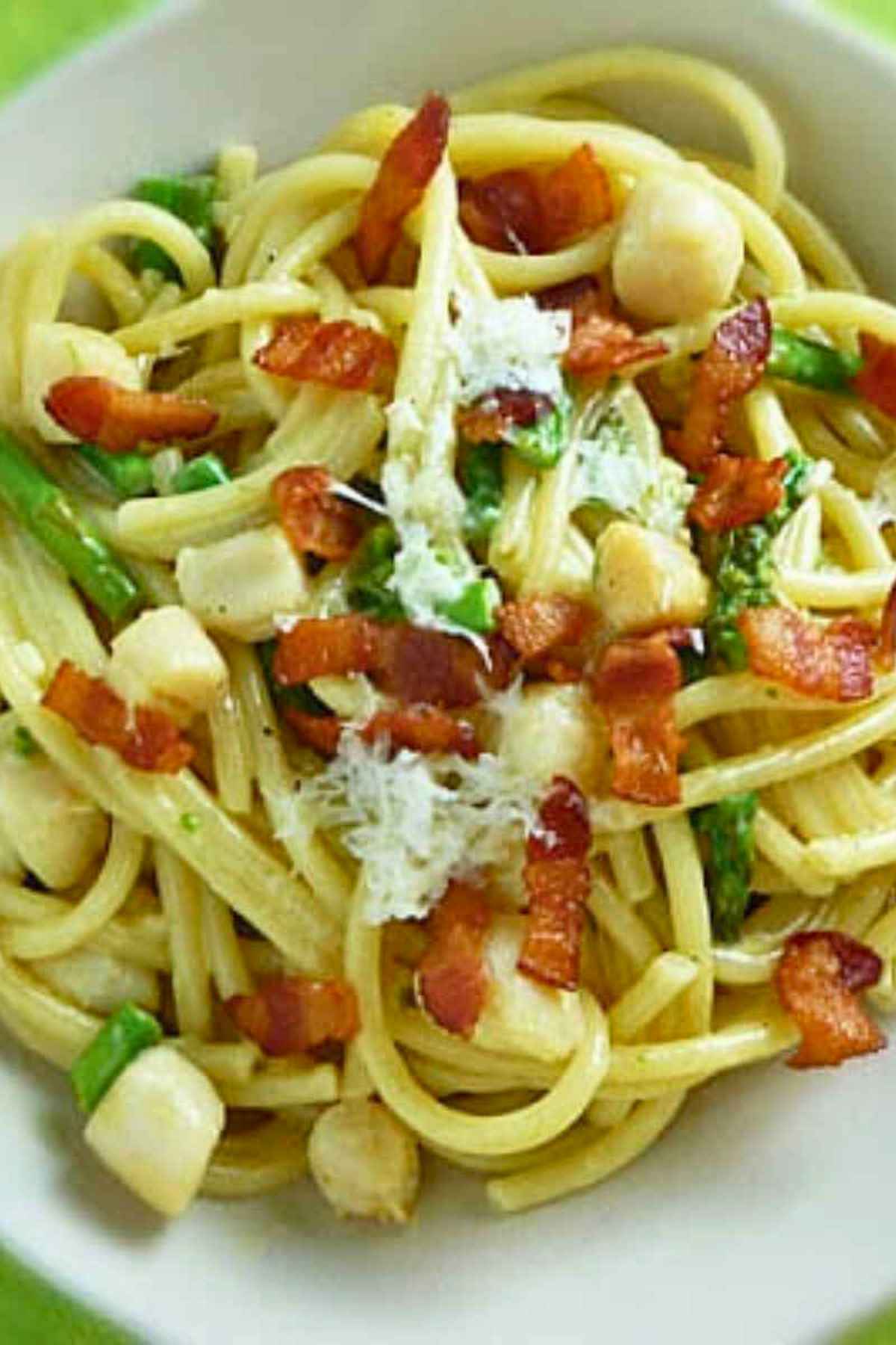 A close-up bowl of creamy scallop carbonara pasta topped with crispy bacon bits, fresh green asparagus pieces, and grated parmesan cheese.