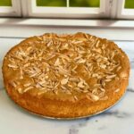 A top-down view of a moist almond flour cake garnished with sliced almonds on a marbled counter top in front of a window.