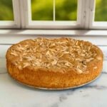 A top-down view of a moist almond flour cake garnished with sliced almonds on a marbled counter top in front of a window.