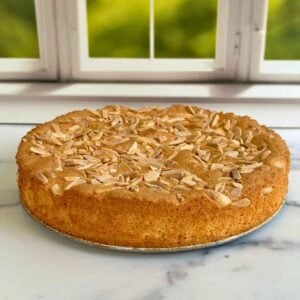 A top-down view of a moist almond flour cake garnished with sliced almonds on a marbled counter top in front of a window.