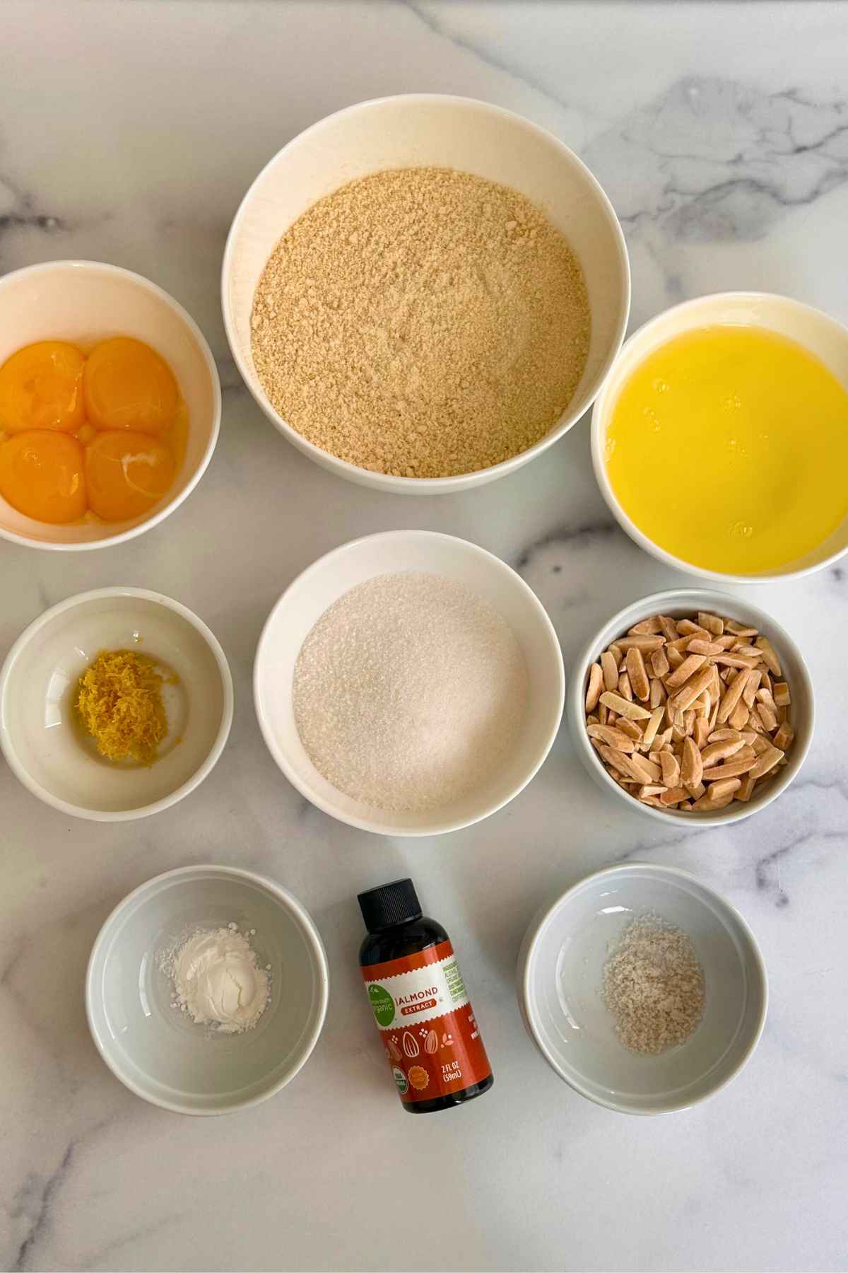 An overhead view of various ingredients arranged in small white bowls on a marble surface, including nut flour, sugar, egg yolks, egg whites, slivered almonds, lemon zest, and a bottle of almond extract.