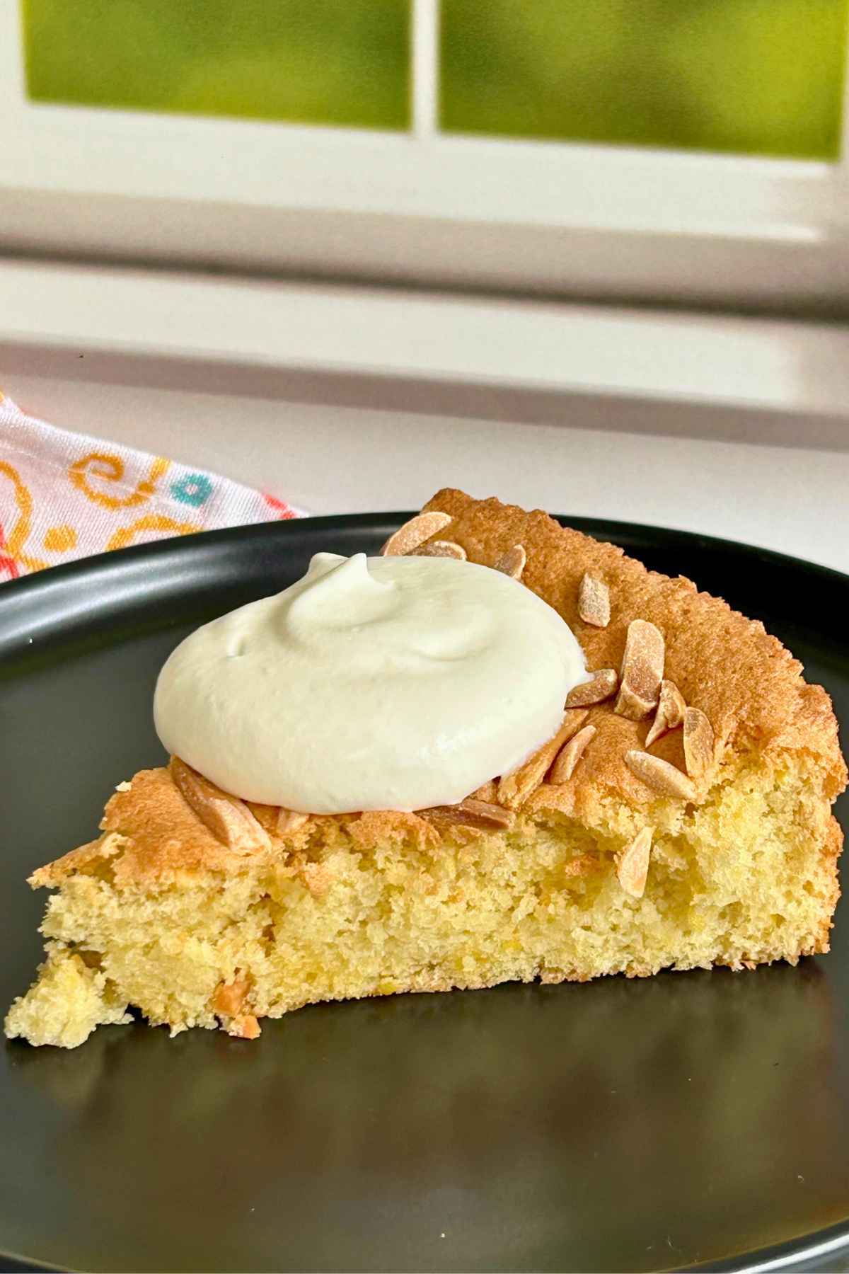 A slice of almond flour cake with whipped cream on a black plate.