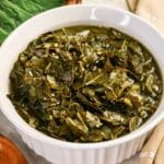 A white fluted bowl filled with tender Southern braised collard greens served in a savory broth with a wooden spoon.
