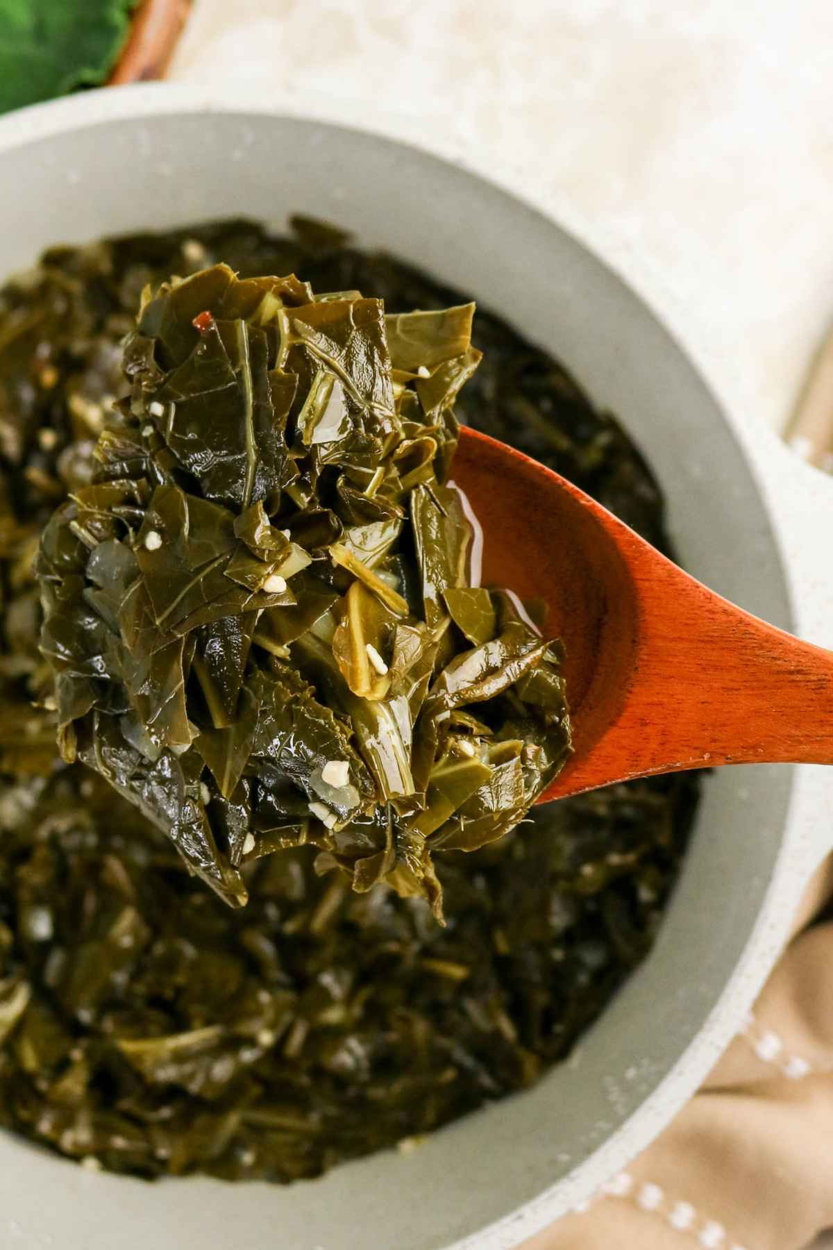 A large wooden spoon lifting a serving of tender braised collard greens from a white cooking pot.