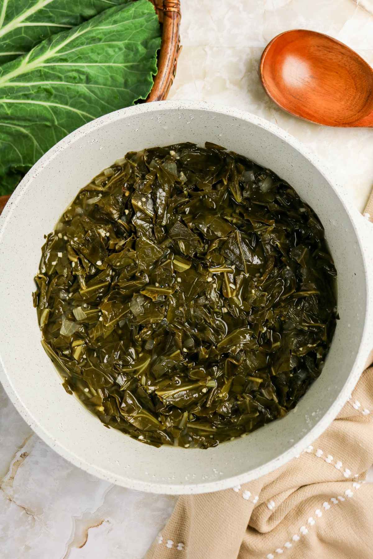 An overhead view of a white bowl filled with tender braised collard greens in a savory broth, served next to a wooden spoon.