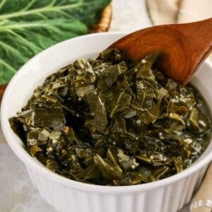 A white fluted bowl filled with tender braised collard greens in a savory broth, shown with a large wooden serving spoon.