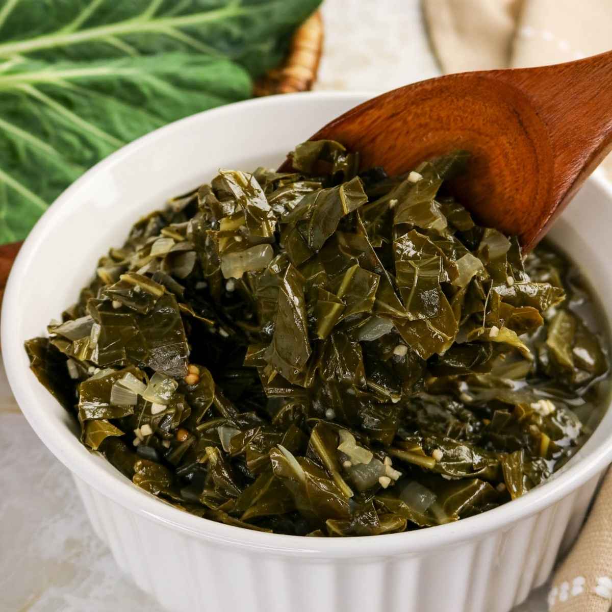 A white fluted bowl filled with tender braised collard greens in a savory broth, shown with a large wooden serving spoon.