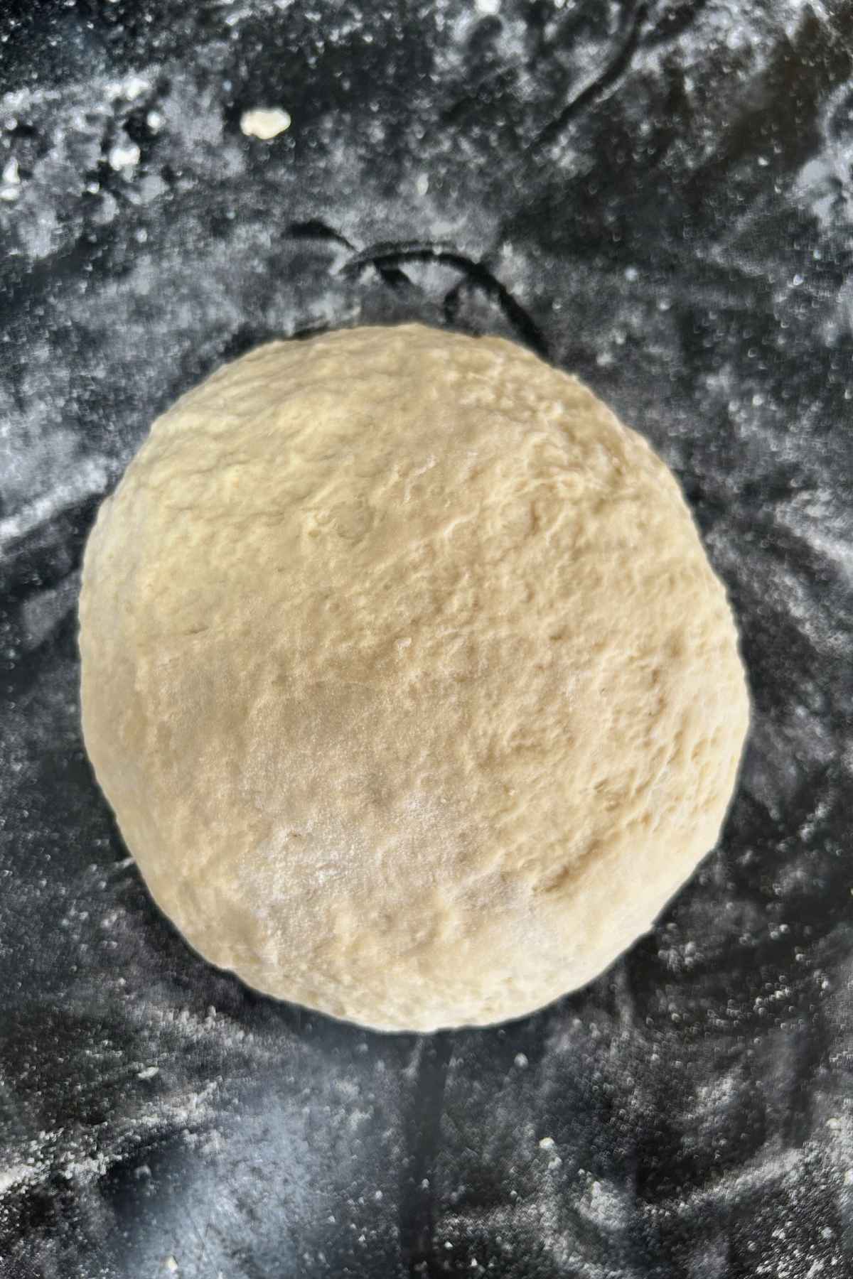 A smooth round ball of dough resting on a dark, floured surface, showing a soft and slightly textured exterior.