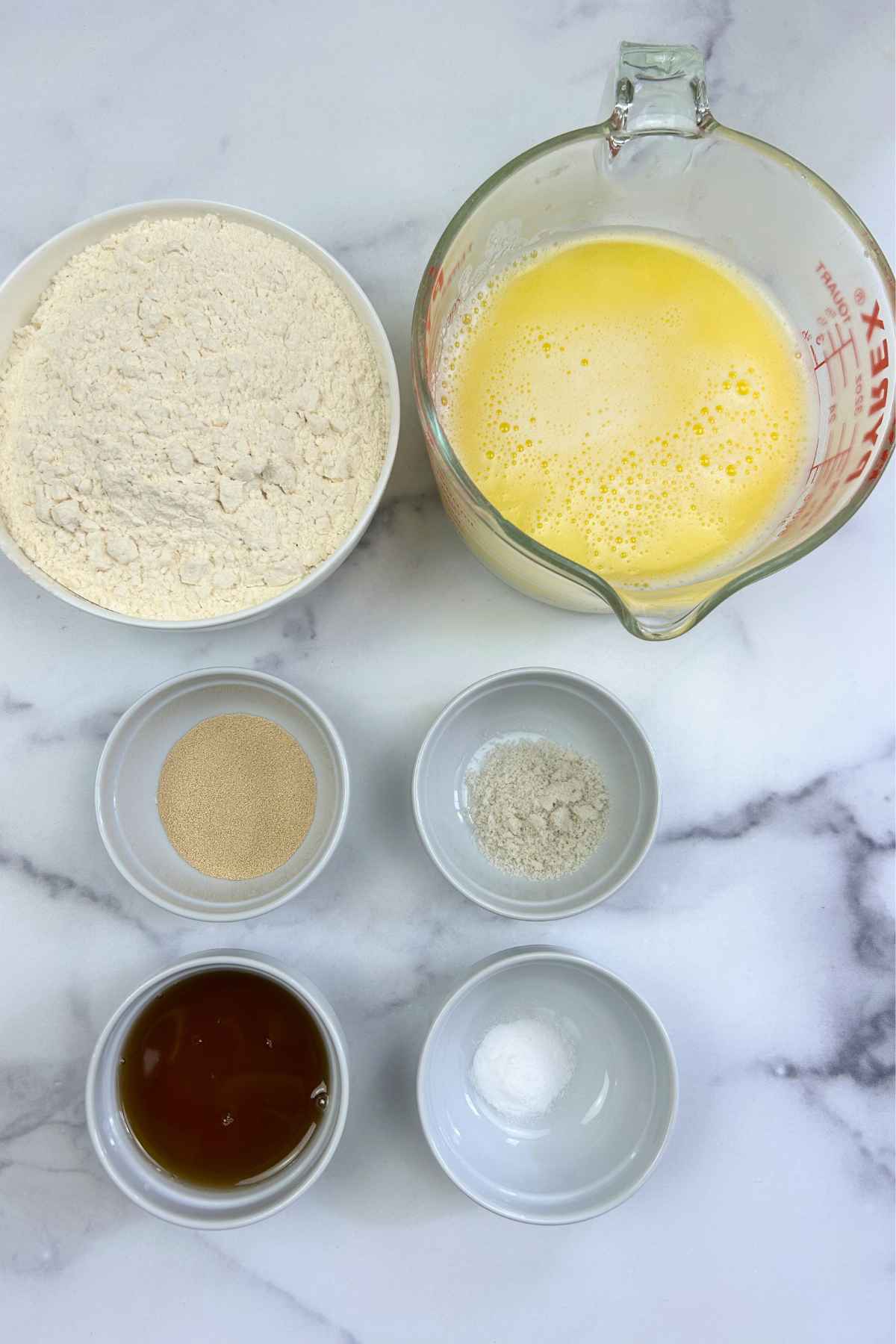 Six glass and ceramic bowls of varying sizes containing flour, a buttermilk and butter mixture, yeast, sugar, salt, and maple syrup arranged on a light colored counter top.