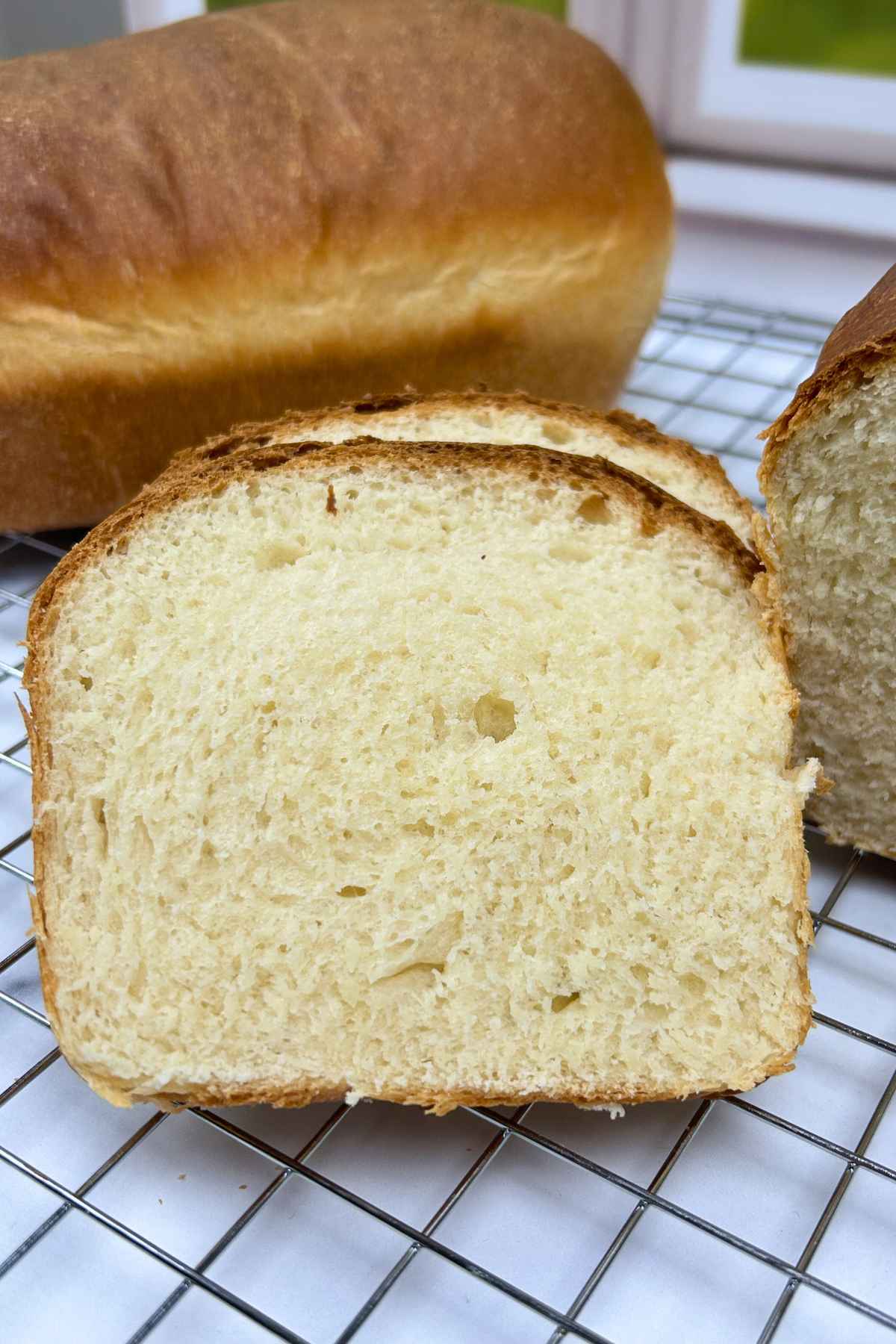 A close up of two thick slices of white homemade bread with a soft, porous interior, leaning against a full loaf on a wire cooling rack.