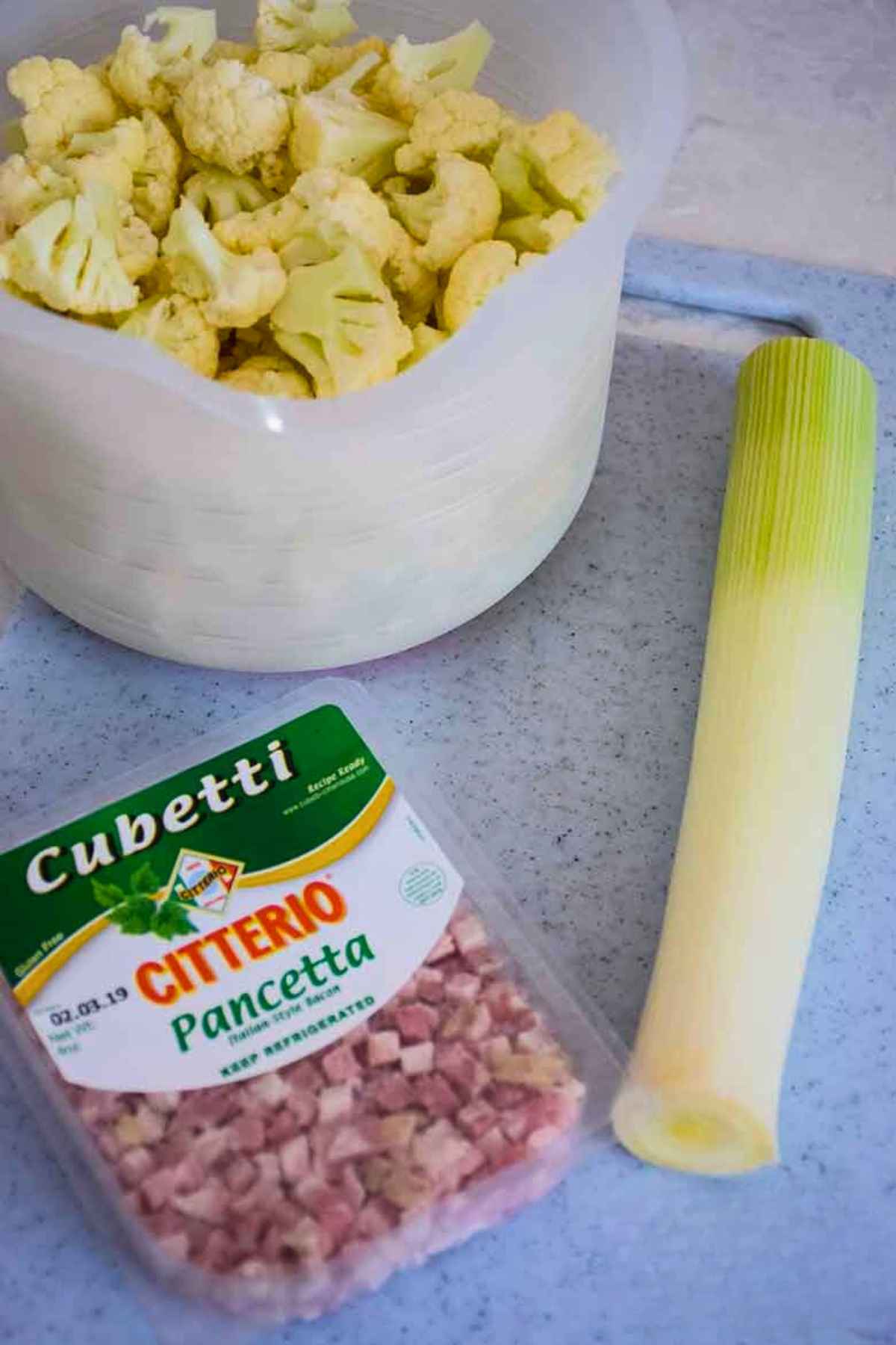 Citterio Pancetta Cubetti packaged, fresh cauliflower florets in a white bowl, and on cleaned and trimmed leek.