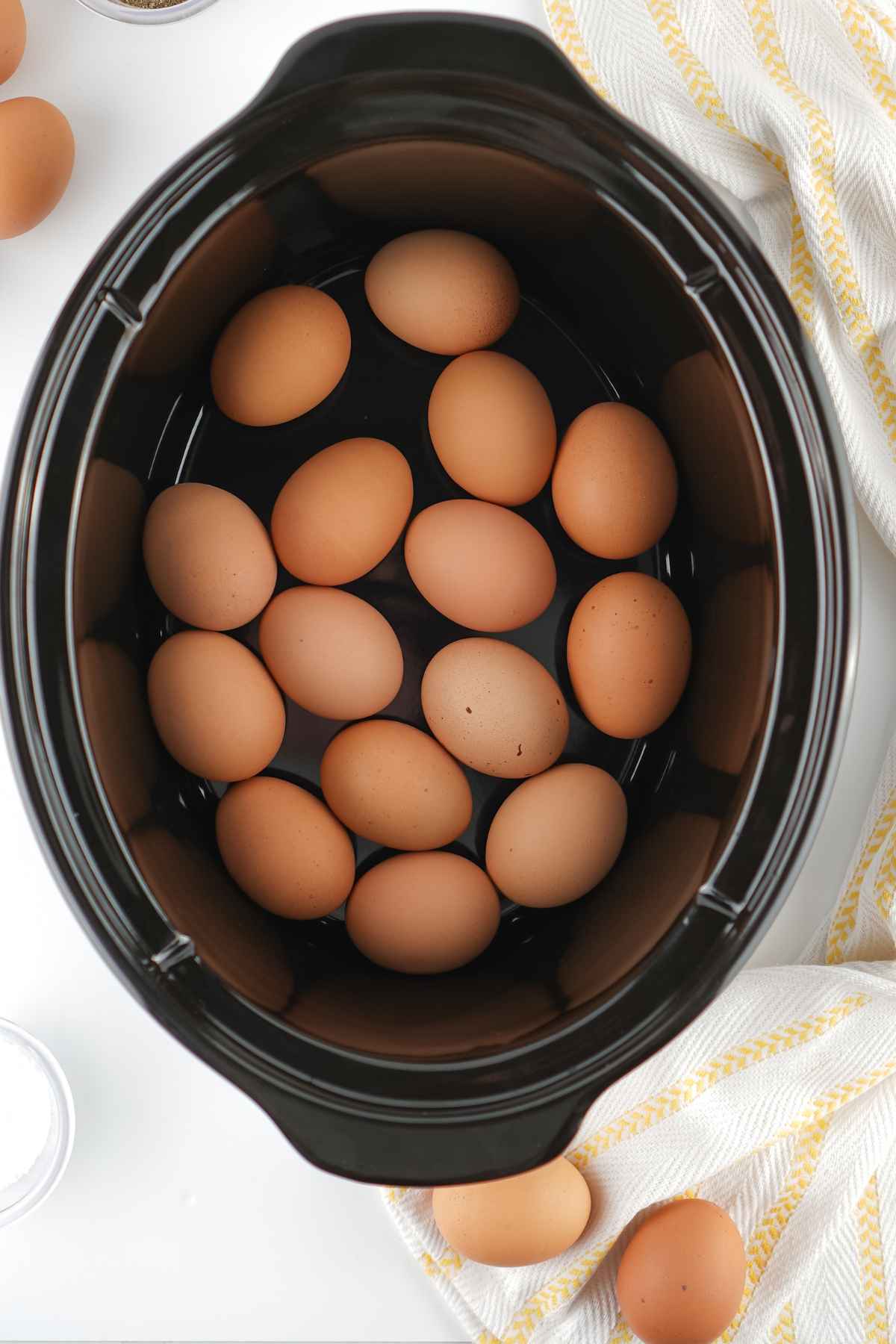 Raw brown eggs placed inside a black ceramic crockpot on a yellow striped kitchen towel.