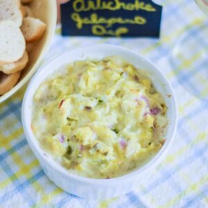 Close-up of a homemade hot Asiago artichoke dip appetizer with a hand-written "artichoke Jalapeno dip" sign in the background.