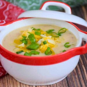 A white bowl with a red rim filled with creamy instant pot cauliflower soup, topped with shredded cheddar cheese and sliced green onions, set against a green and white patterned napkin.