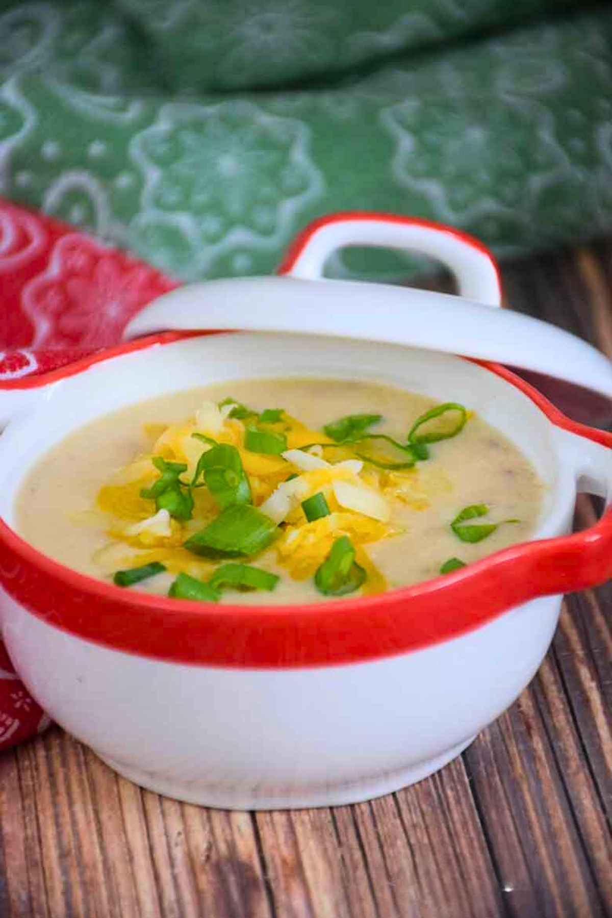 A white bowl with a red rim filled with creamy instant pot cauliflower soup, topped with shredded cheddar cheese and sliced green onions, set against a green and white patterned napkin.