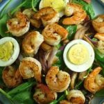 A vibrant Shrimp Spinach Salad featuring sauteed shrimp, sliced hard-boiled eggs, and caramelized onions served in a blue bowl with a warm bacon dressing.