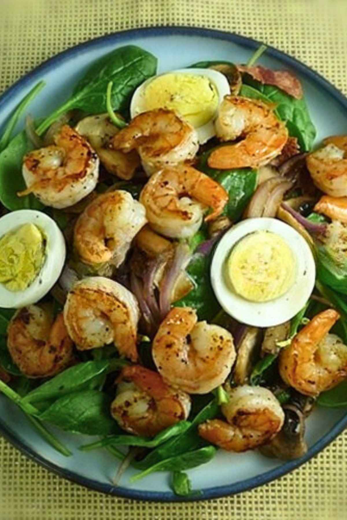 A vibrant Shrimp Spinach Salad featuring sauteed shrimp, sliced hard-boiled eggs, and caramelized onions served in a blue bowl with a warm bacon dressing.