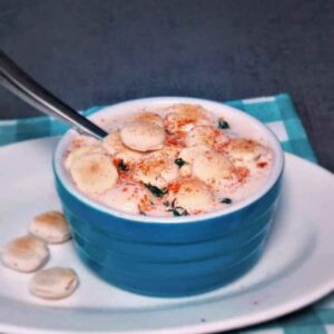A blue ceramic bowl filled with creamy slow cooker clam chowder topped with several oyster crackers and a sprinkle of paprika, served with a spoon on a blue checkered napkin.
